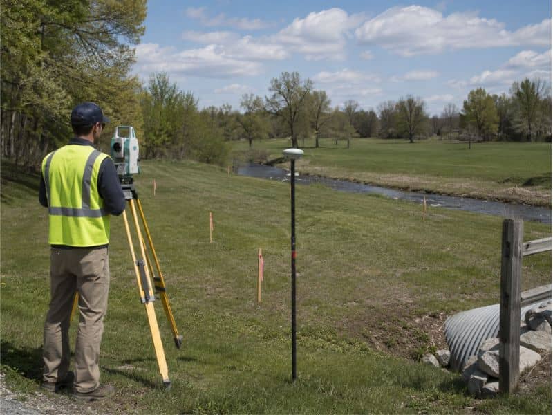 Surveyor in a high-visibility vest uses a tripod-mounted instrument to measure land by a river in a park.