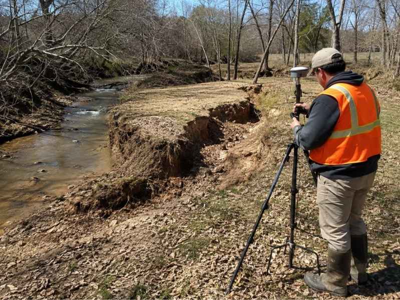 Worker in an orange safety vest sets up a tripod-mounted surveying instrument by an eroded riverbank, measuring the ground